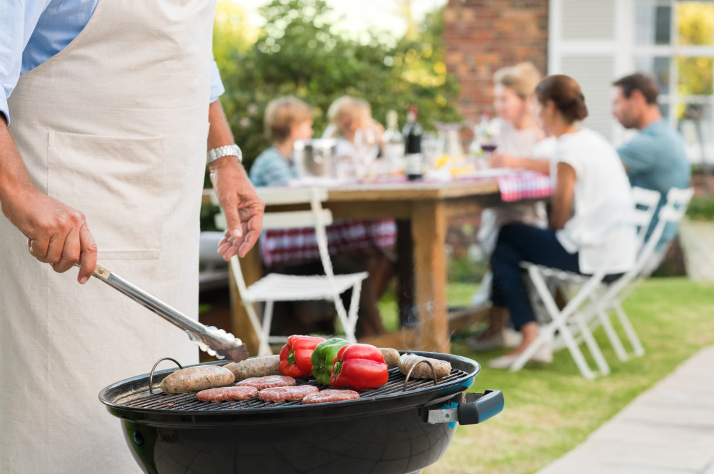 Man cooking food on an outdoor grill with guests seated in the background.