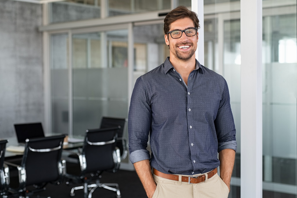 Young bespectacled man smiling while standing outside a conference room.